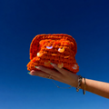 Mini clutch Ibiza orange avec des sequins oranges, tenu par une main avec le ciel bleu en arrière plan