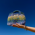 Sac Havana bleu, lilas, jaune et vert, tenu par une main avec le ciel bleu en arrière plan