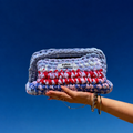 Clutch Azur rose, bleu, rouge et blanc, tenu par une main avec le ciel bleu en arrière plan.