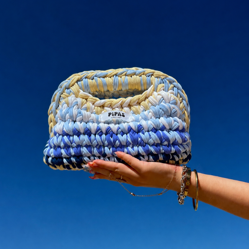 Clutch Capri bleu, jaune et blanc tenu par une main avec le ciel bleu en arrière plan