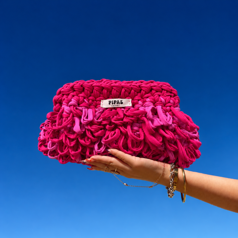 Clutch Medusa rose fushia bouclé, tenu par une main avec le ciel bleu en fond