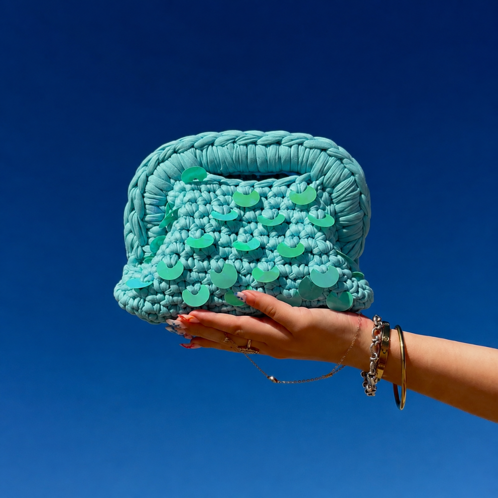 Mini Clutch Palma turquoise avec sequins turquoises, tenu par une main avec le ciel bleu en fond