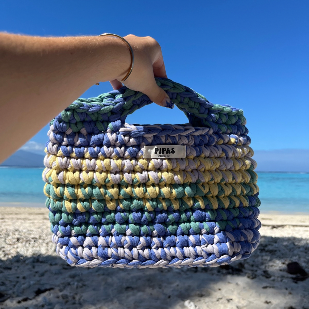 Sac Havana bleu, jaune, lilas et vert, tenu par une main à la plage