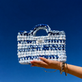 Sac Mykonos bleu et blanc, tenu par une main avec le ciel bleu en arrière plan