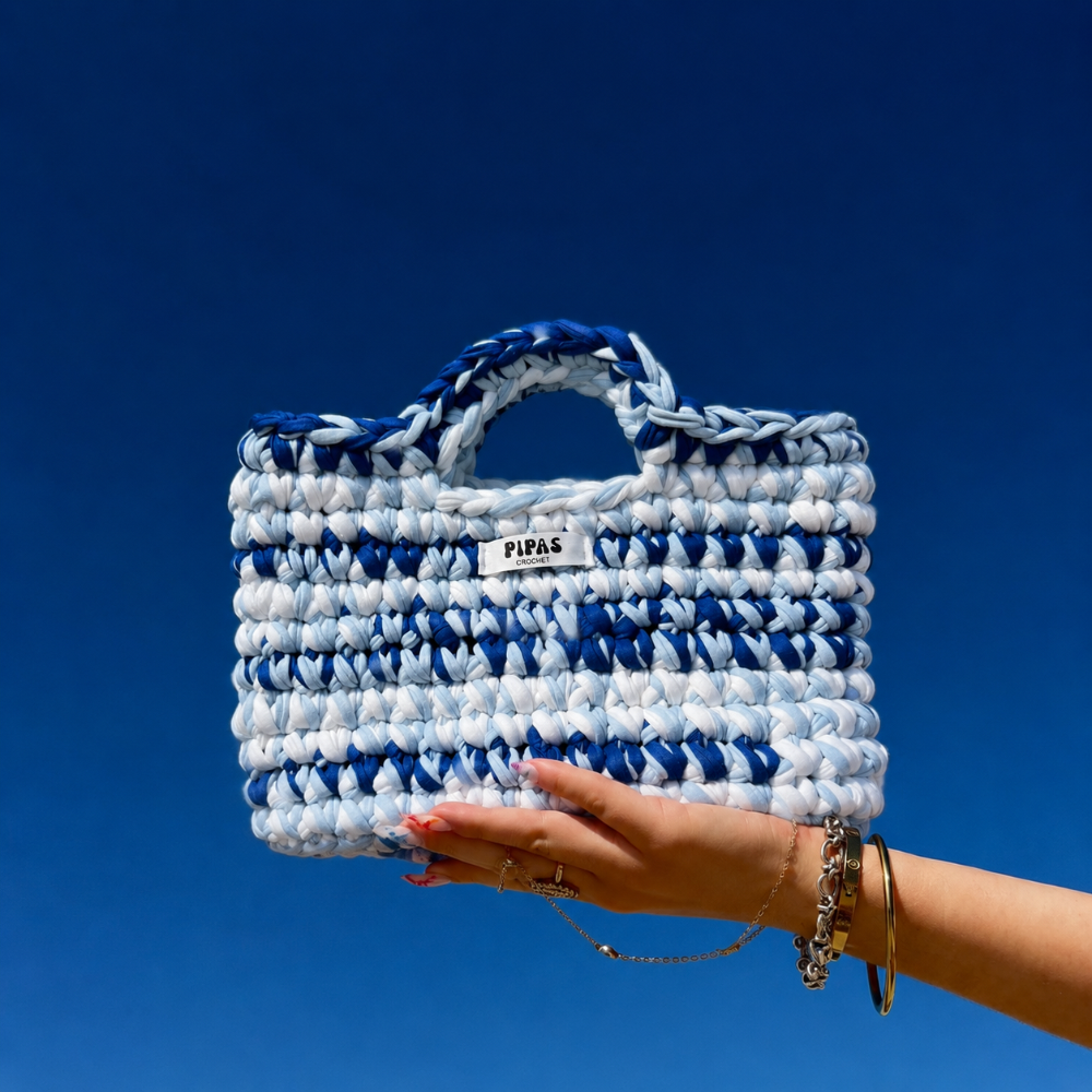 Sac Mykonos bleu et blanc, tenu par une main avec le ciel bleu en arrière plan