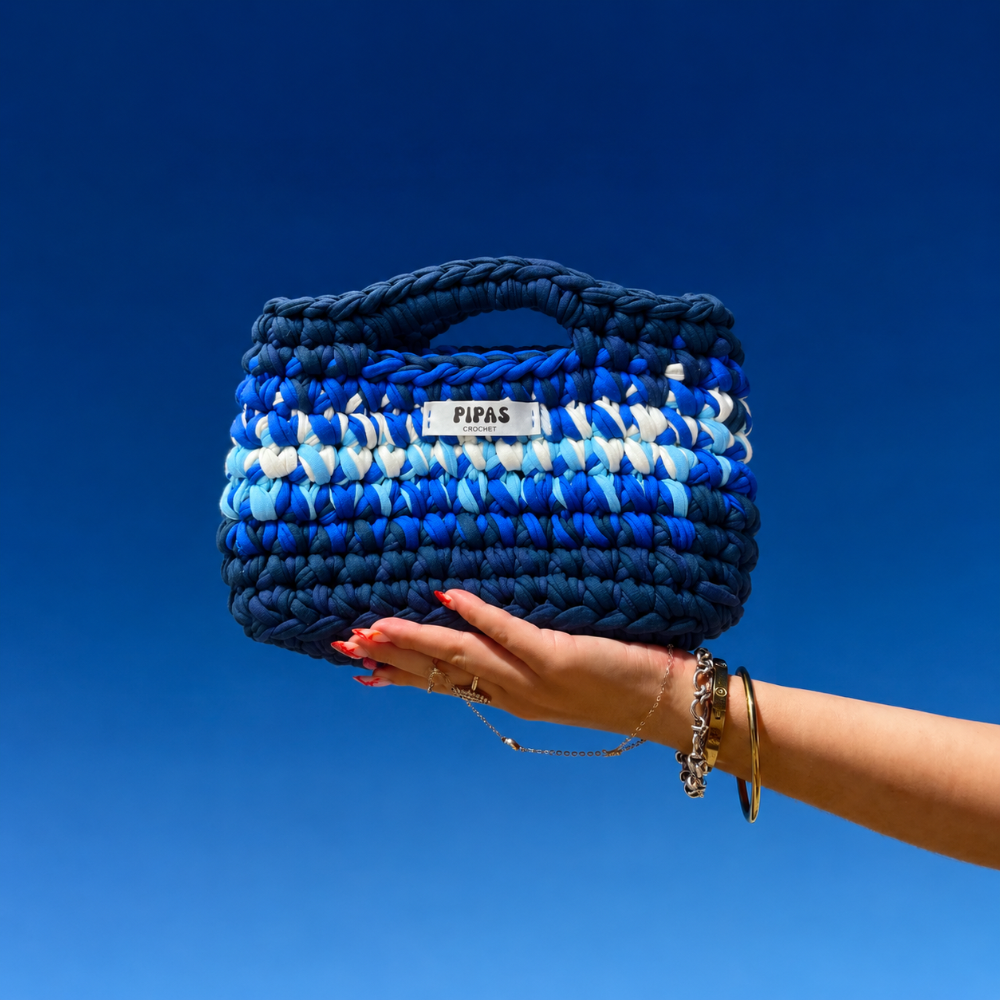 Sac Santorini bleu et blanc, tenu par une main avec le ciel bleu en fond