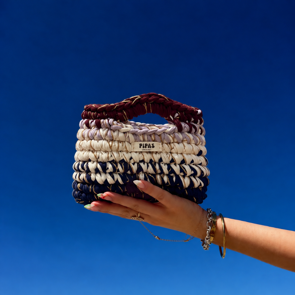 Sac Toscane bordeaux, bleu marine, lilas, blanc avec du fil doré tenu par une main avec le ciel bleu en arrière plan
